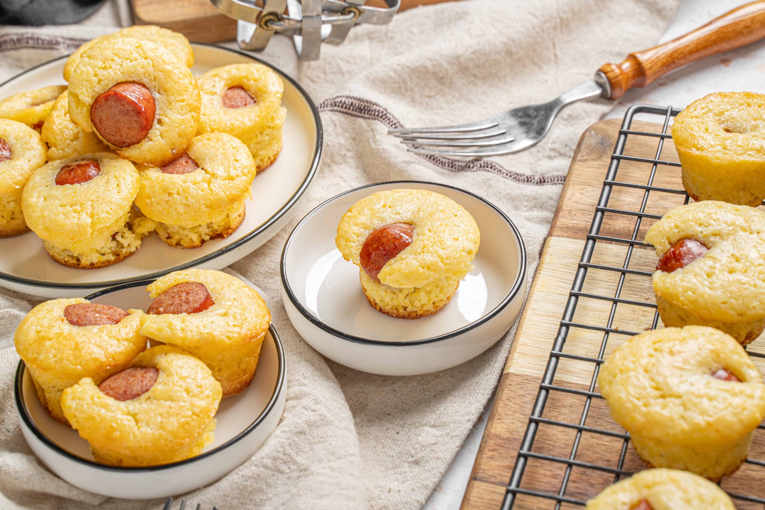 Fluffy sausage and cornbread muffins served on white plates, with a wire rack and wooden cutting board for cooling, set on a rustic beige tablecloth. Perfect for breakfast or brunch recipes.