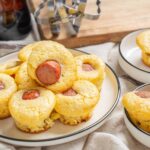 Homemade mini cornbread muffins filled with sliced sausage, served on white and black-rimmed plates, with a rustic wooden background and bottle of syrup in the background.