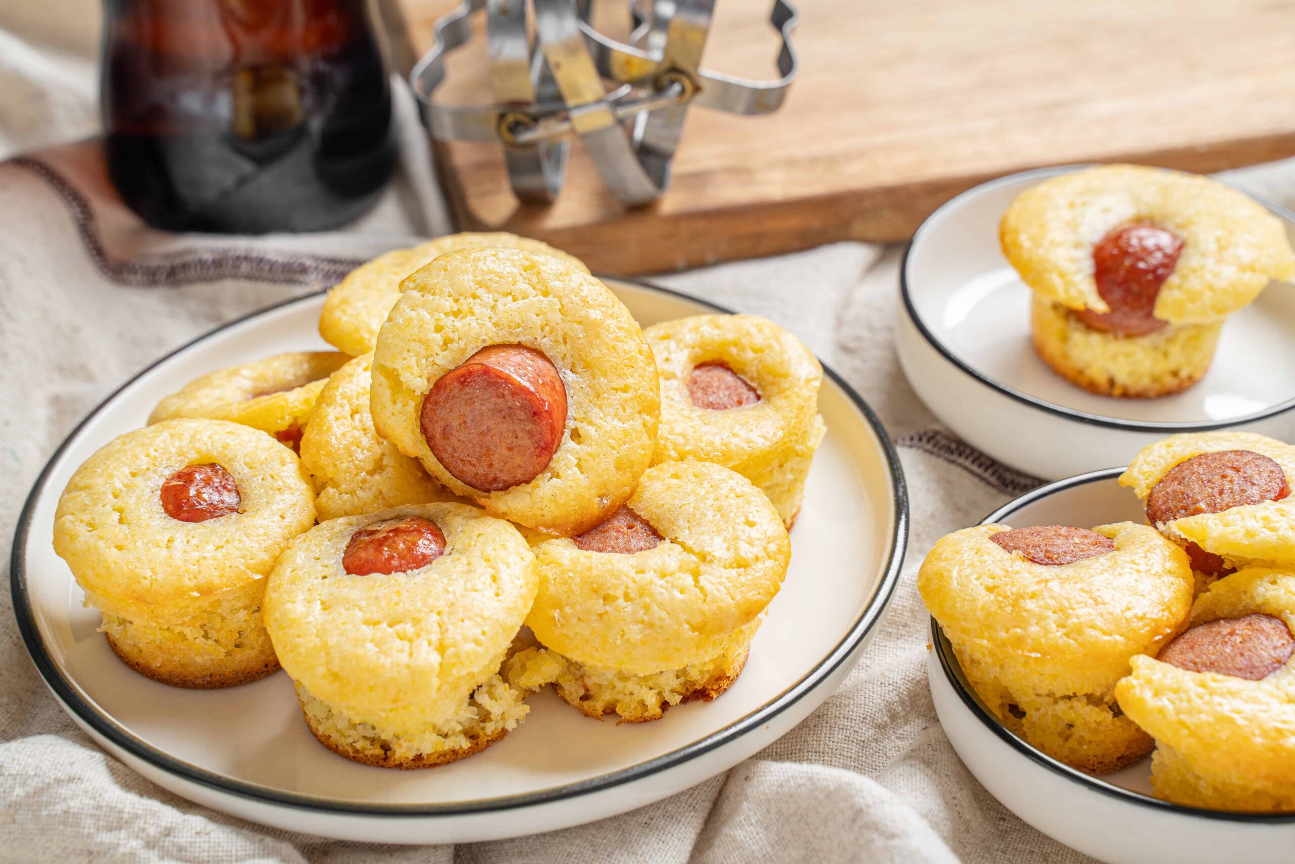 Homemade mini cornbread muffins filled with sliced sausage, served on white and black-rimmed plates, with a rustic wooden background and bottle of syrup in the background.