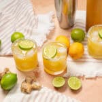 Refreshing citrus ginger lemonade with lime and lemon slices, ice, and salted rim, served in glass on pink textured background with fresh limes, lemons, ginger, and drink ingredients for summer cocktail.