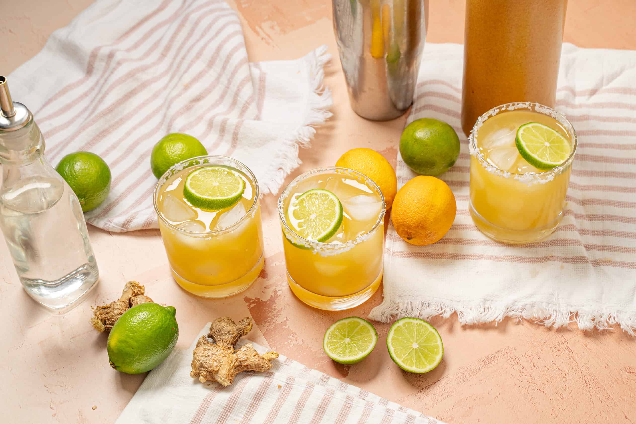 Refreshing citrus ginger lemonade with lime and lemon slices, ice, and salted rim, served in glass on pink textured background with fresh limes, lemons, ginger, and drink ingredients for summer cocktail.
