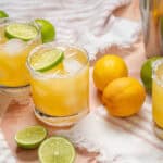 Freshly prepared margarita cocktails with ice, lime wedges, and salted rims on a light pink table, surrounded by fresh lemons and limes for a refreshing summer drink.