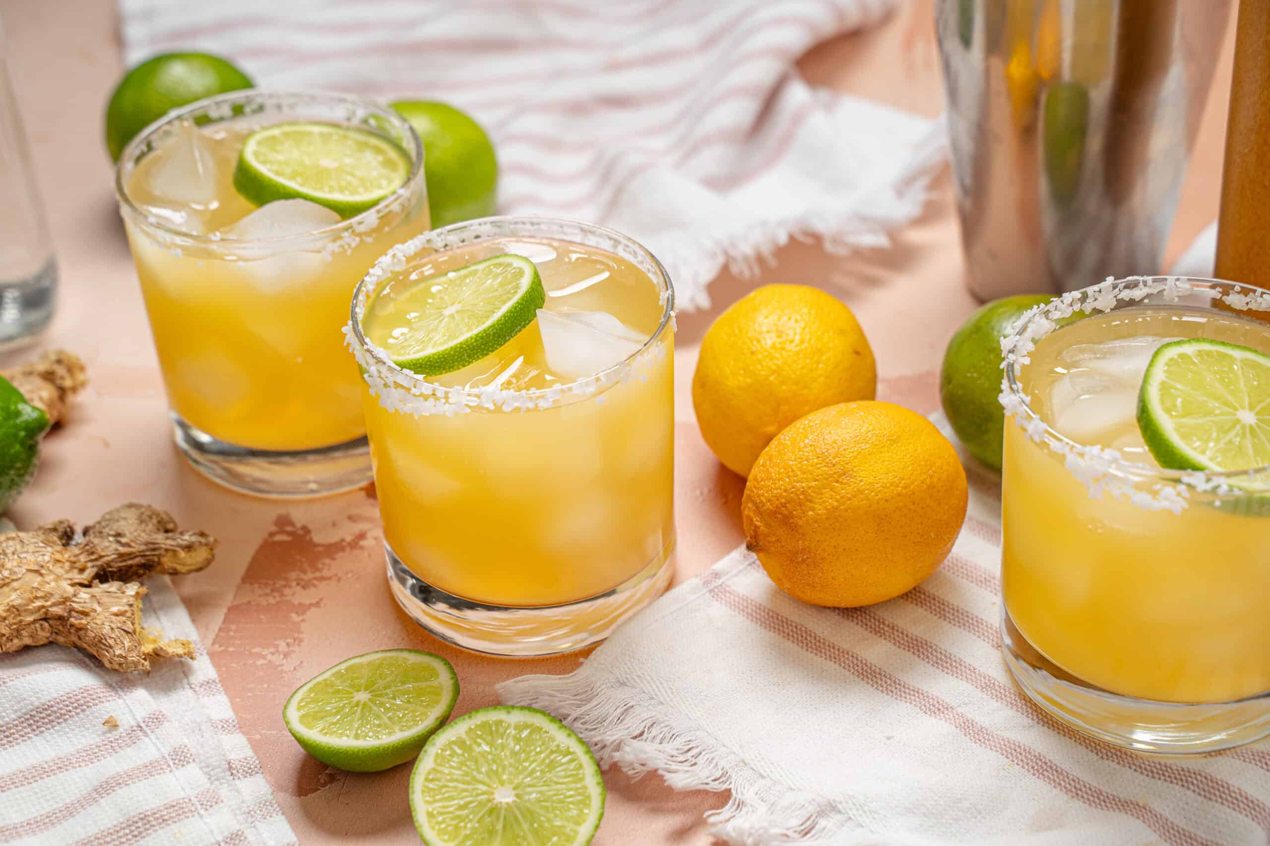 Freshly prepared margarita cocktails with ice, lime wedges, and salted rims on a light pink table, surrounded by fresh lemons and limes for a refreshing summer drink.