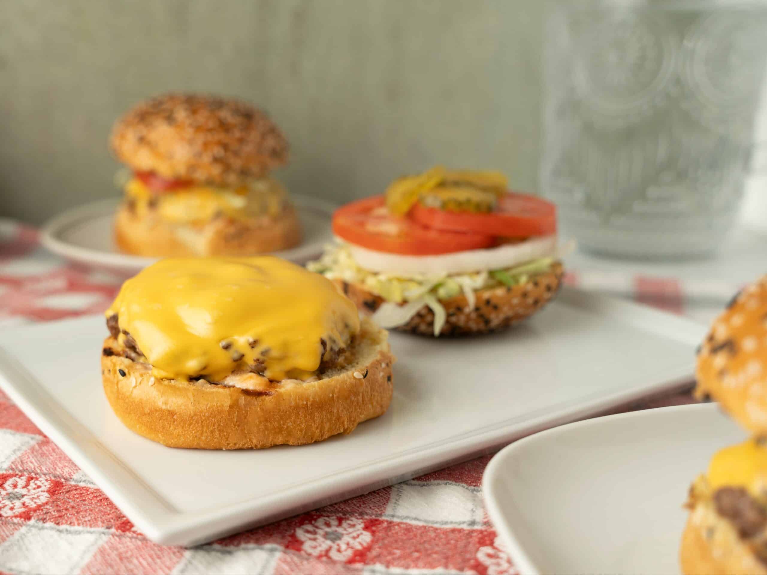 Cheeseburger sliders with melted cheese, fresh tomato, lettuce, pickles on sesame seed bun on white tray, delicious homemade mini cheeseburgers for lunch or dinner, tasty fast food meal, crispy and juicy patties, appetizing snack, savory American style burger, gourmet slider sandwiches, mouthwatering cheeseburger bites, popular comfort food, quick and easy to prepare.