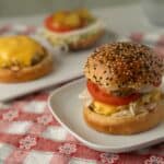 Golden sesame seed burger bun with fresh tomato, lettuce, pickles, cheese, and grilled chicken on a white plate, with additional cheeseburgers in the background, served on a red checkered tablecloth.