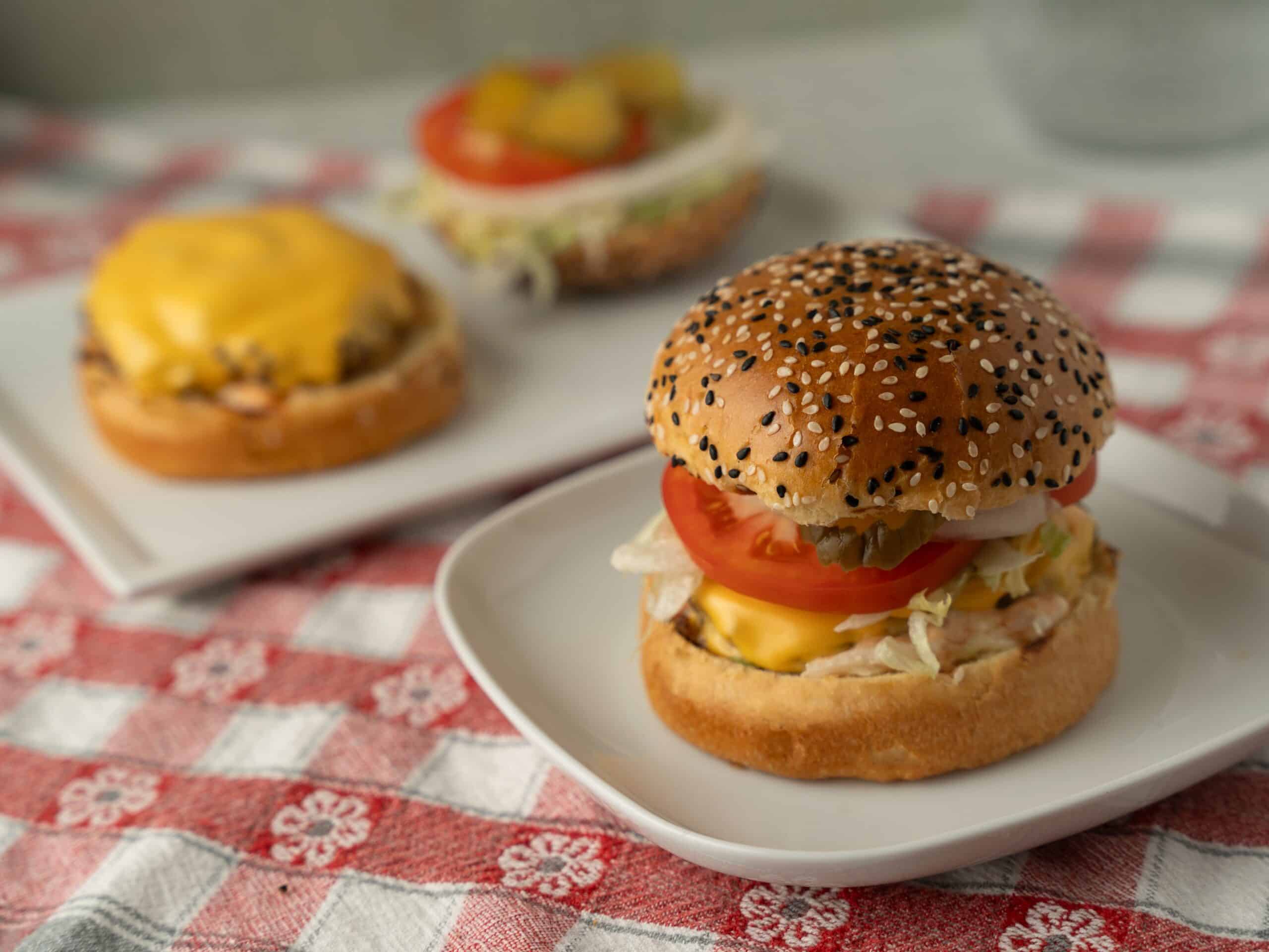 Golden sesame seed burger bun with fresh tomato, lettuce, pickles, cheese, and grilled chicken on a white plate, with additional cheeseburgers in the background, served on a red checkered tablecloth.