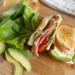 Fresh homemade sandwich with avocado, tomato, lettuce, ham, and creamy spread on toasted bread. Perfect for a healthy lunch or snack, served on a wooden cutting board with slices of avocado and cherry tomato.
