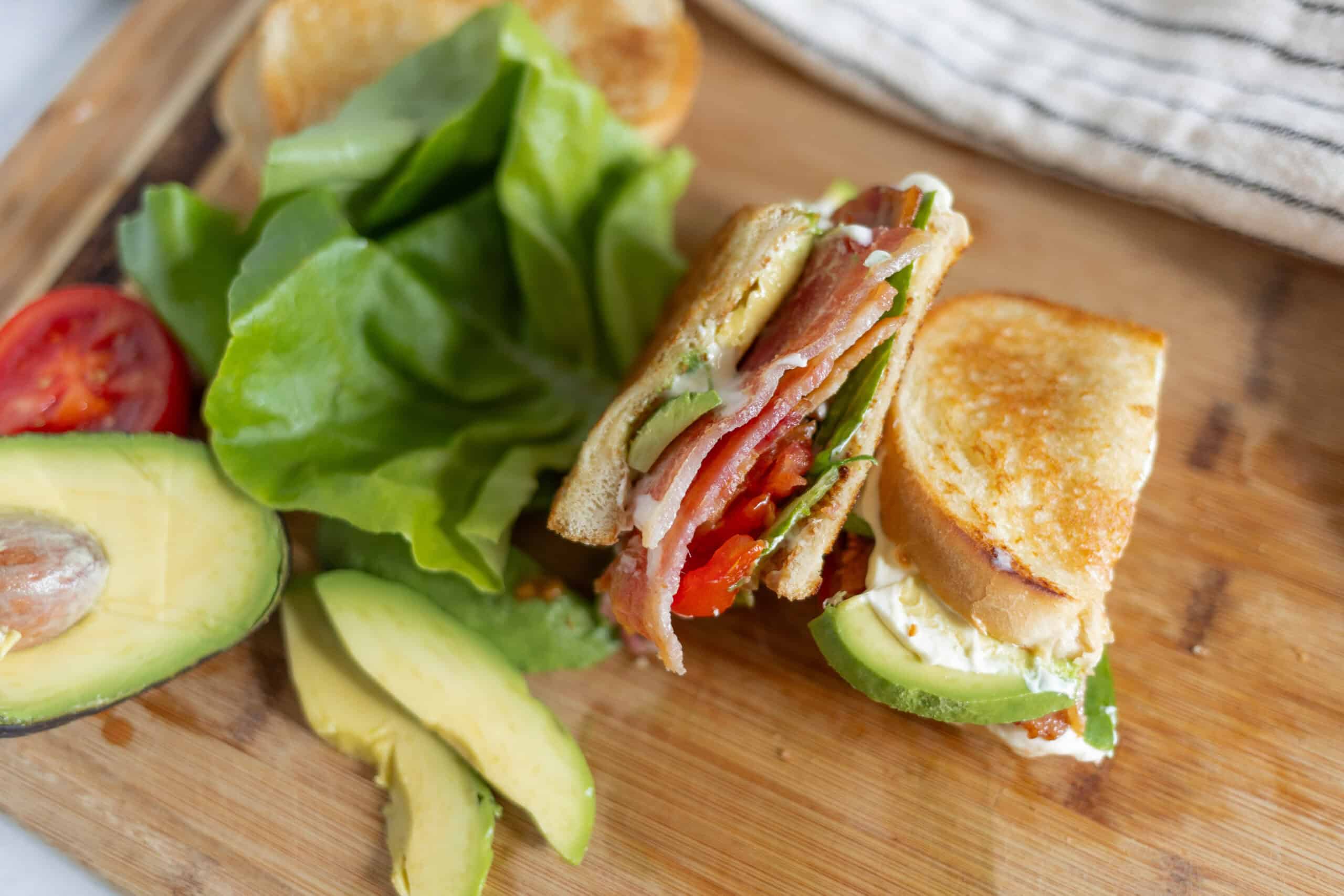 Fresh homemade sandwich with avocado, tomato, lettuce, ham, and creamy spread on toasted bread. Perfect for a healthy lunch or snack, served on a wooden cutting board with slices of avocado and cherry tomato.