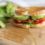 Savory breakfast bacon, avocado, tomato, and lettuce sandwich on a toasted bagel, served with fresh sliced avocado on a wooden cutting board.
