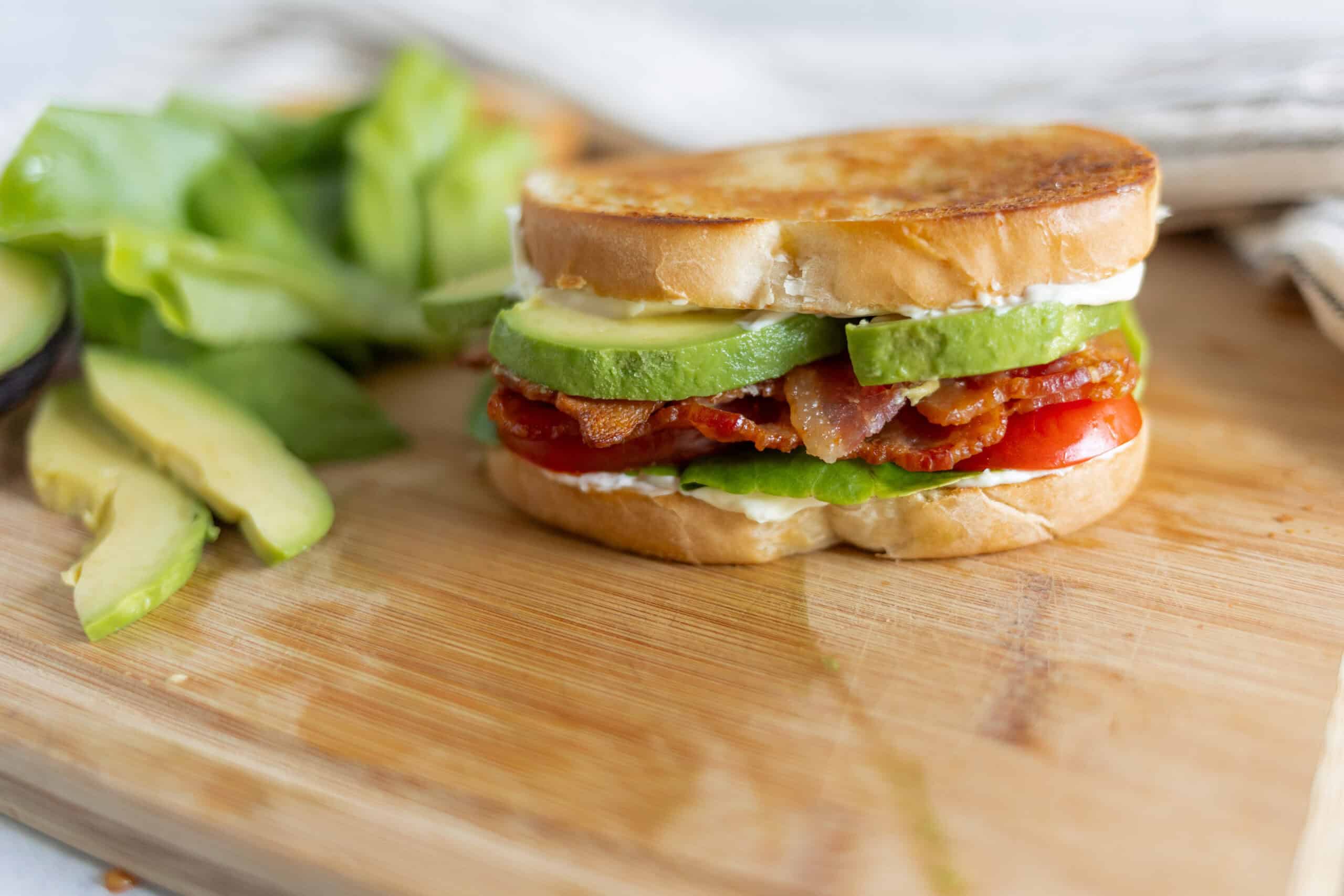 Savory breakfast bacon, avocado, tomato, and lettuce sandwich on a toasted bagel, served with fresh sliced avocado on a wooden cutting board.