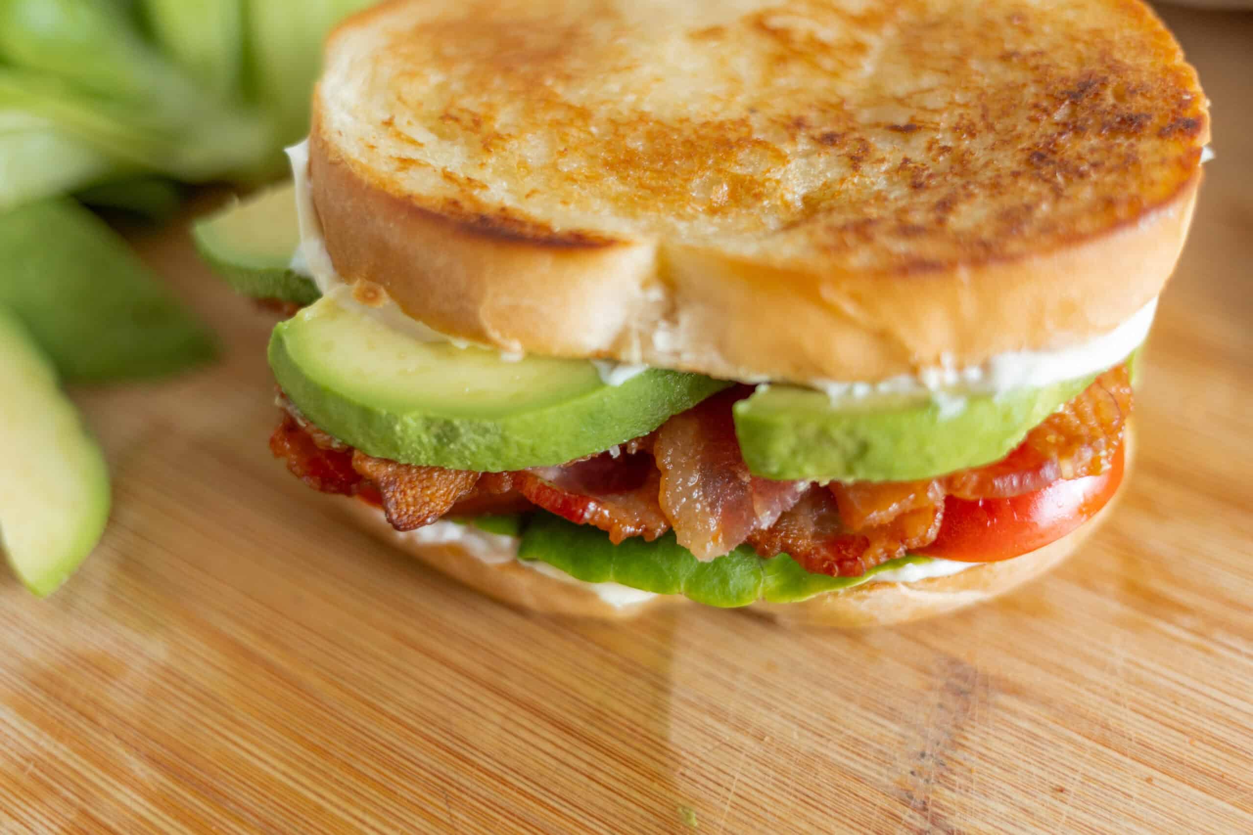 Crispy bacon and fresh avocado slices in a toasted bread sandwich on a wooden cutting board. Delicious homemade BLT sandwich with crispy bacon, ripe tomatoes, and creamy avocado.