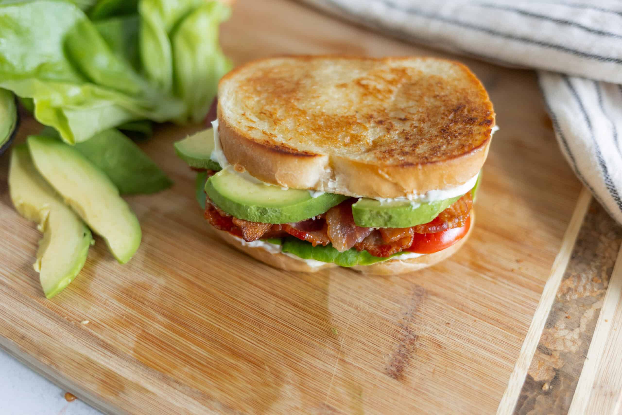 Fresh homemade BLT sandwich with crispy bacon, ripe tomato, creamy mayonnaise, crunchy lettuce, and sliced avocado on toasted bread on a wooden cutting board.