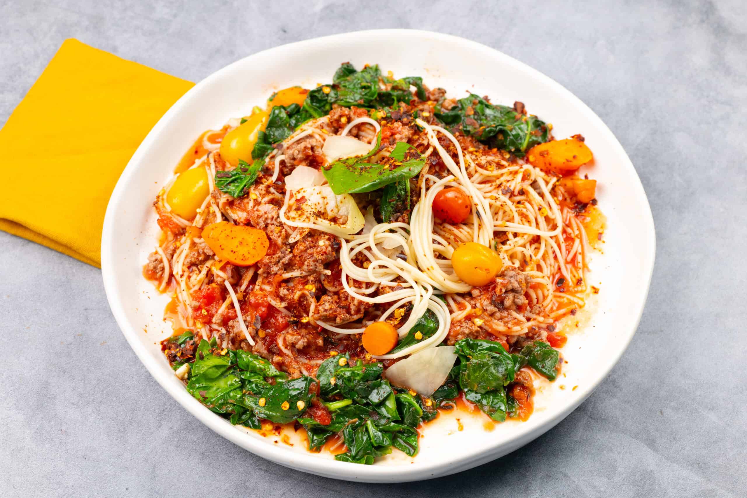 Spaghetti Bolognese with fresh vegetables and herbs on a white plate, served with a yellow napkin, showcasing a flavorful and colorful pasta dish.