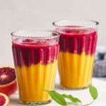 Vibrant two-tone fruit smoothies in tall glasses with layered red and yellow colors, surrounded by blood oranges and green leaves on a white surface for refreshing, healthy drink imagery.