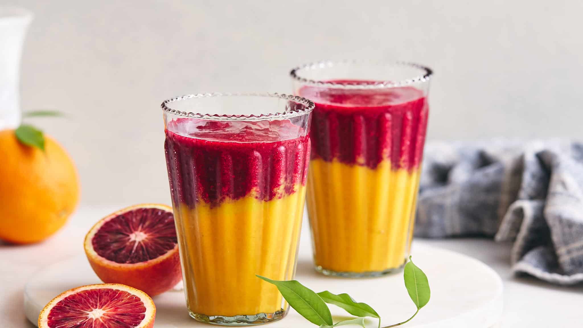 Vibrant two-tone fruit smoothies in tall glasses with layered red and yellow colors, surrounded by blood oranges and green leaves on a white surface for refreshing, healthy drink imagery.