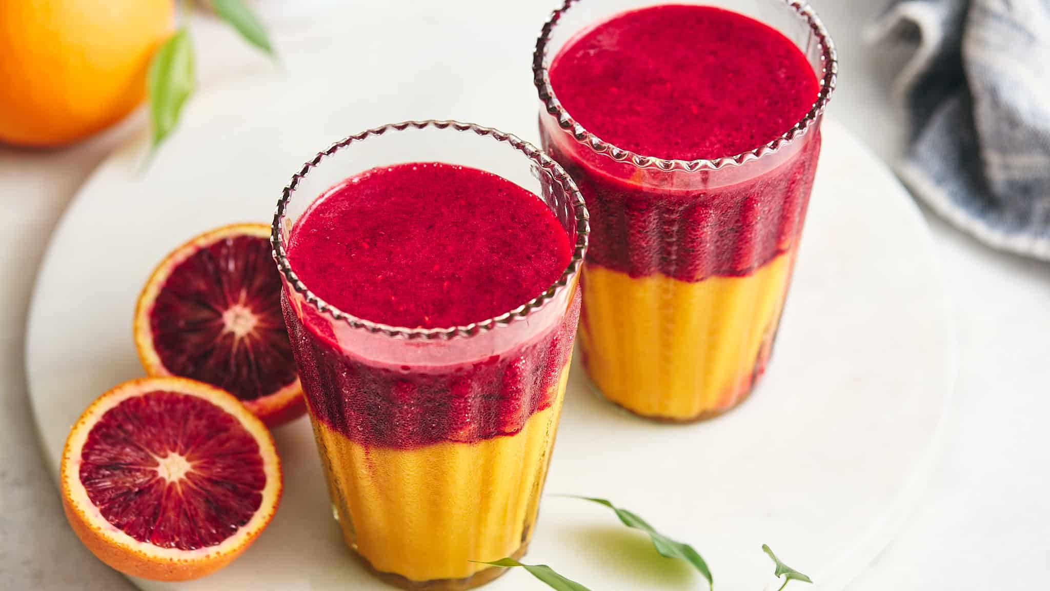 Fresh blood orange and mango smoothies served in tall glasses, garnished with blood orange slices, on a white plate. A vibrant, healthy smoothie recipe perfect for breakfast or summer refreshment.