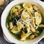 Fresh spinach and white beans stuffed pasta served in a white bowl with shredded cheese on top, garnished with chopped herbs, on a marble surface with a gray striped cloth.
