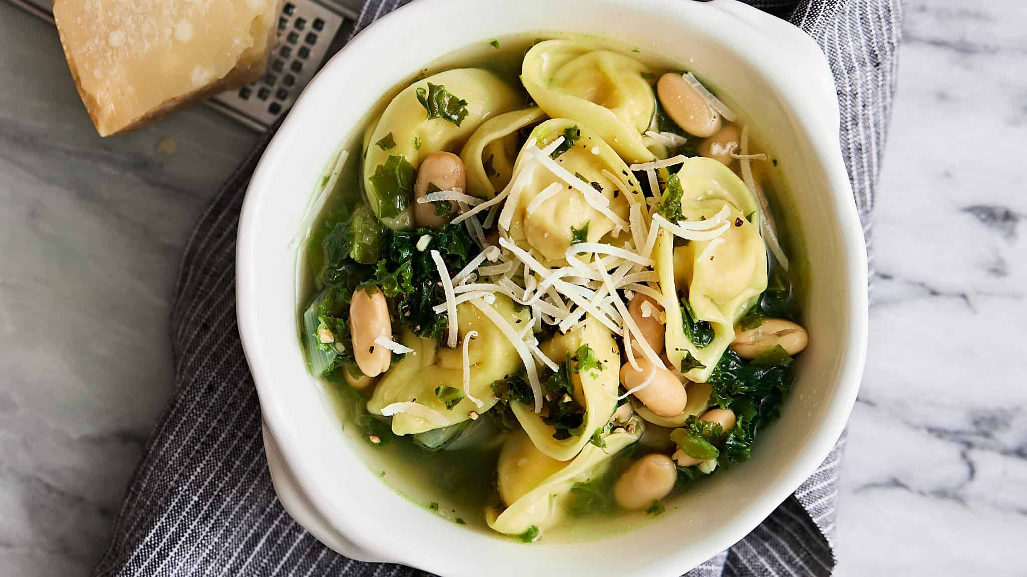 Fresh spinach and white beans stuffed pasta served in a white bowl with shredded cheese on top, garnished with chopped herbs, on a marble surface with a gray striped cloth.