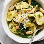 Fresh tortellini salad with white beans, kale, and shredded cheese in a white bowl. Made with spinach, herbs, and olive oil, perfect for light lunches or dinner.