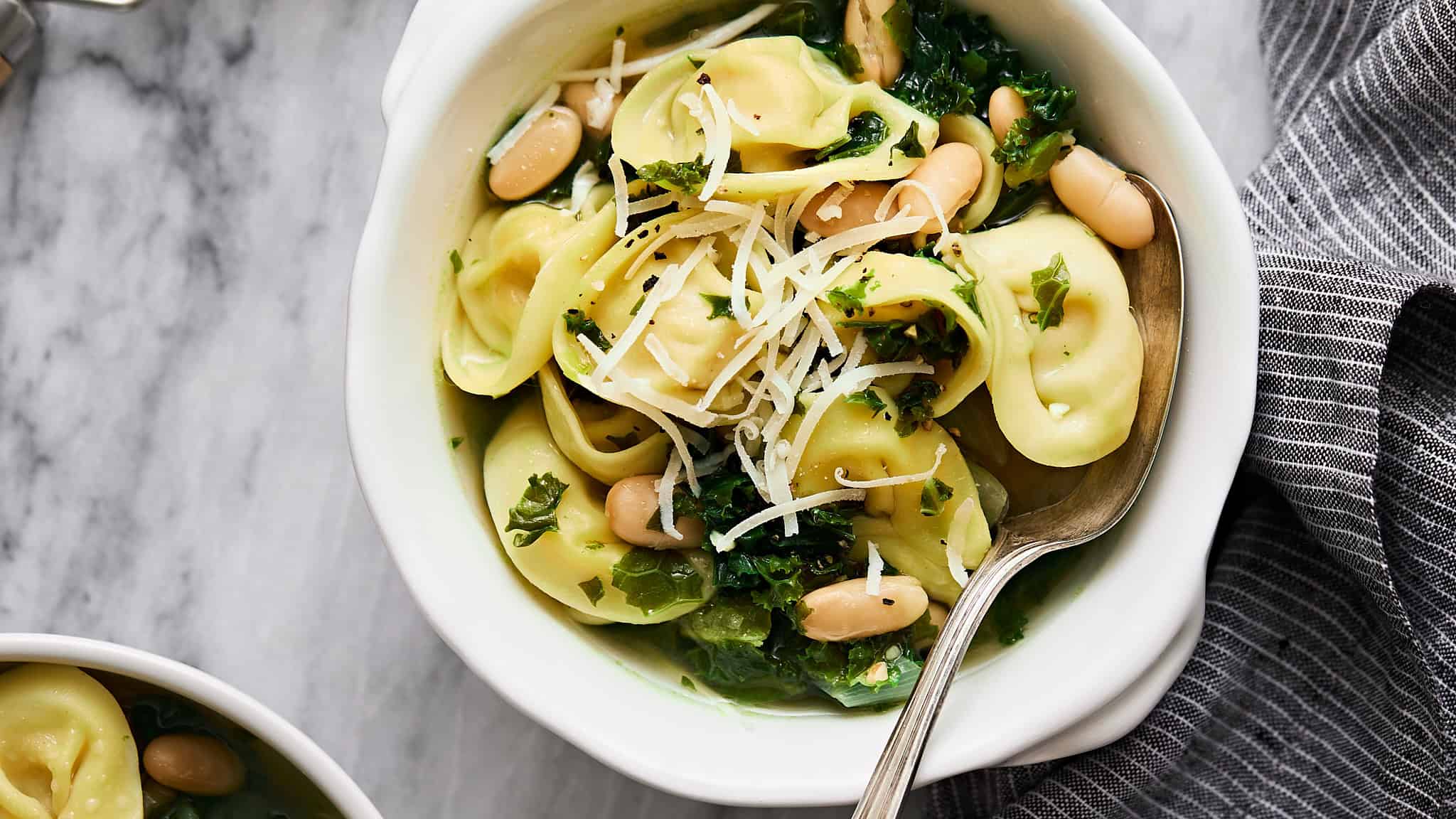 Fresh tortellini salad with white beans, kale, and shredded cheese in a white bowl. Made with spinach, herbs, and olive oil, perfect for light lunches or dinner.
