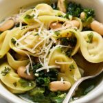 Creamy tortellini salad with white beans, kale, and shredded cheese in a bowl, garnished with herbs and served with a spoon. Perfect for quick, healthy meals.