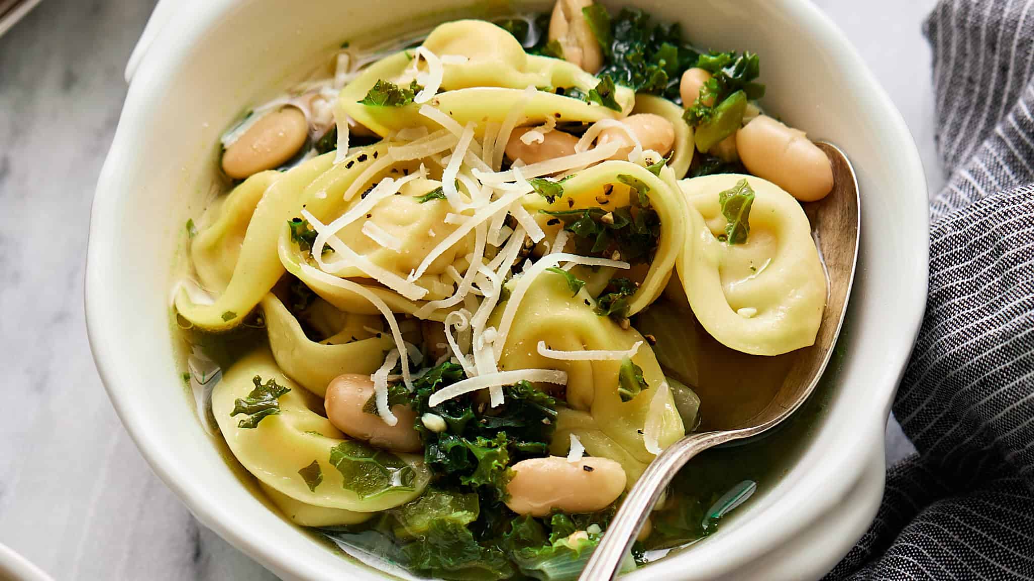 Creamy tortellini salad with white beans, kale, and shredded cheese in a bowl, garnished with herbs and served with a spoon. Perfect for quick, healthy meals.