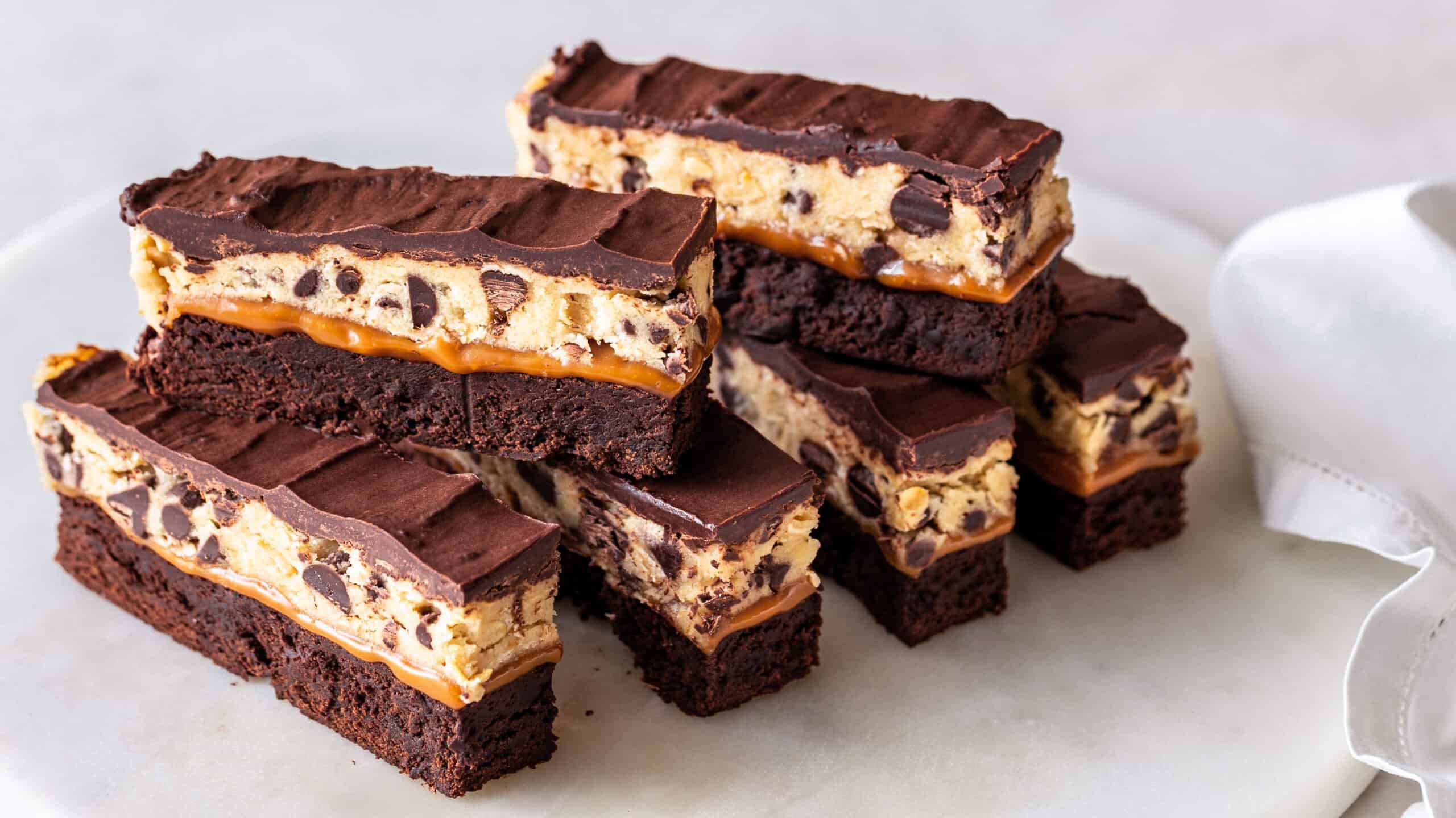 Rich chocolate layered cookie bars with caramel, marshmallow, and chocolate chips on a white plate, perfect for dessert or sweet treat.