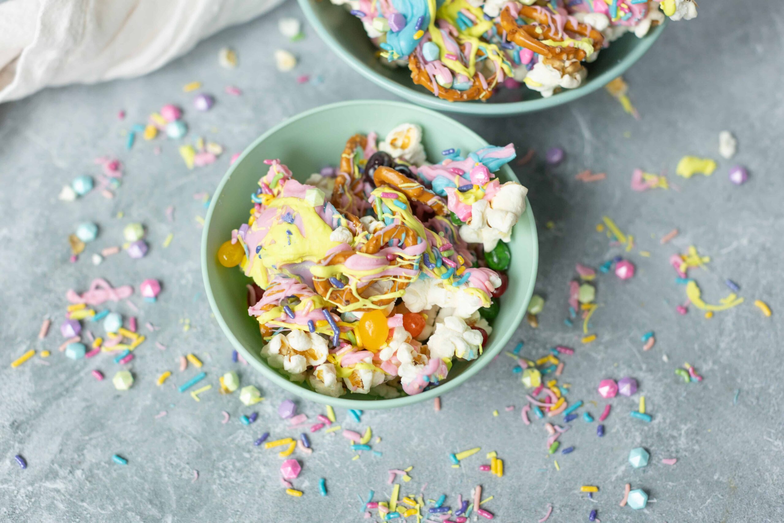 Colorful rainbow popcorn with sprinkles and drizzle in pastel colors served in green bowls on a textured gray surface.