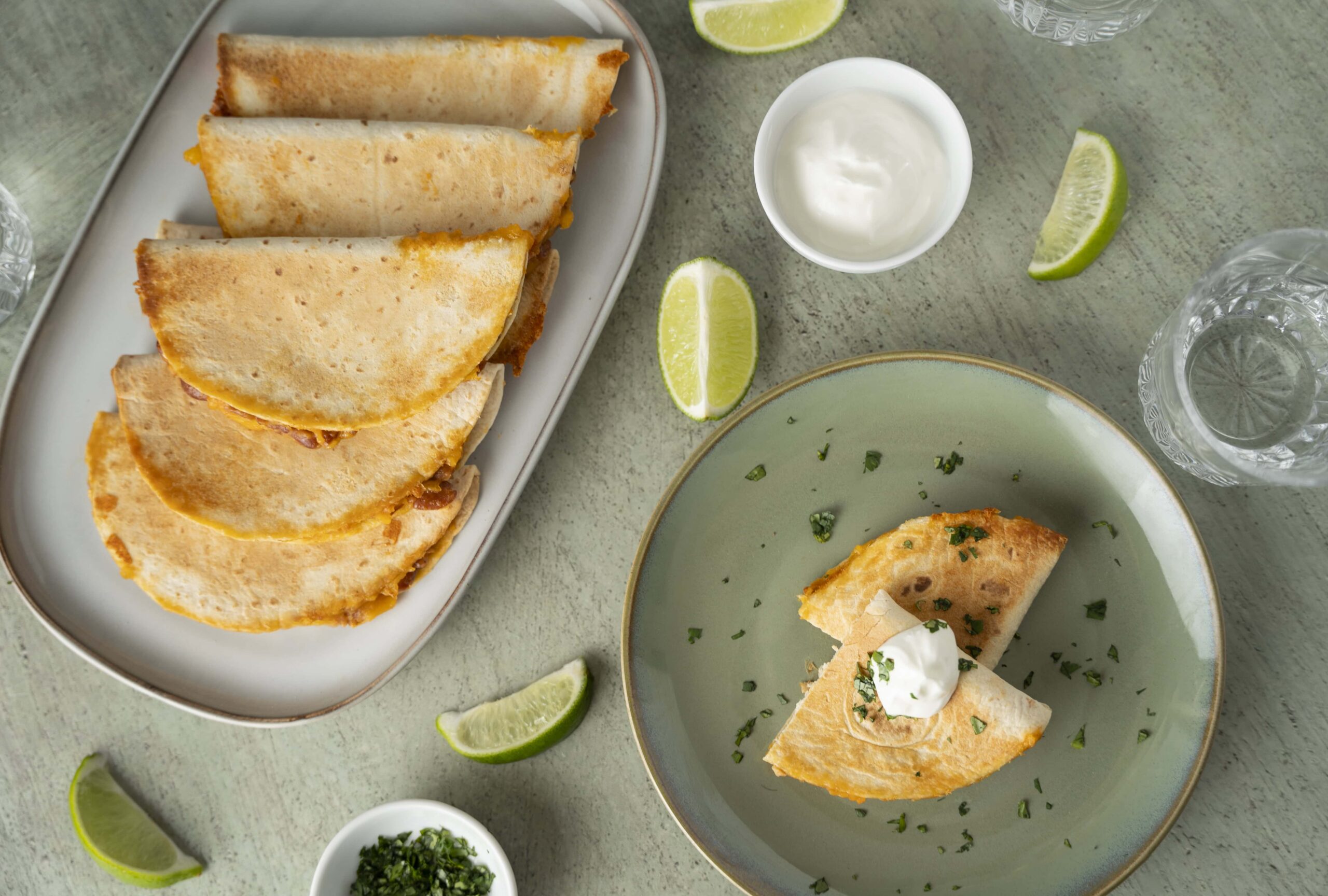 Golden cheese quesadillas with melted cheese and crispy tortilla served with lime wedges, sour cream, and fresh herbs on a light green textured table.