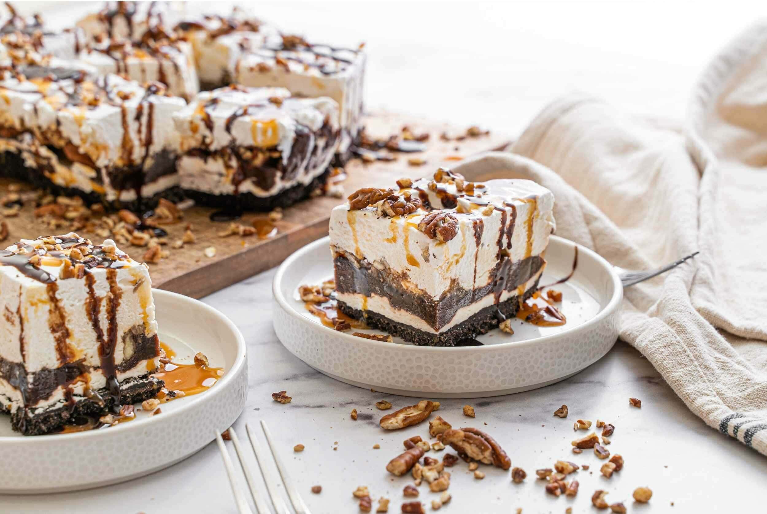 Delicious layered ice cream dessert with chocolate chunks, caramel drizzle, and chopped nuts on top, served on white plates with a whole cake in the background.