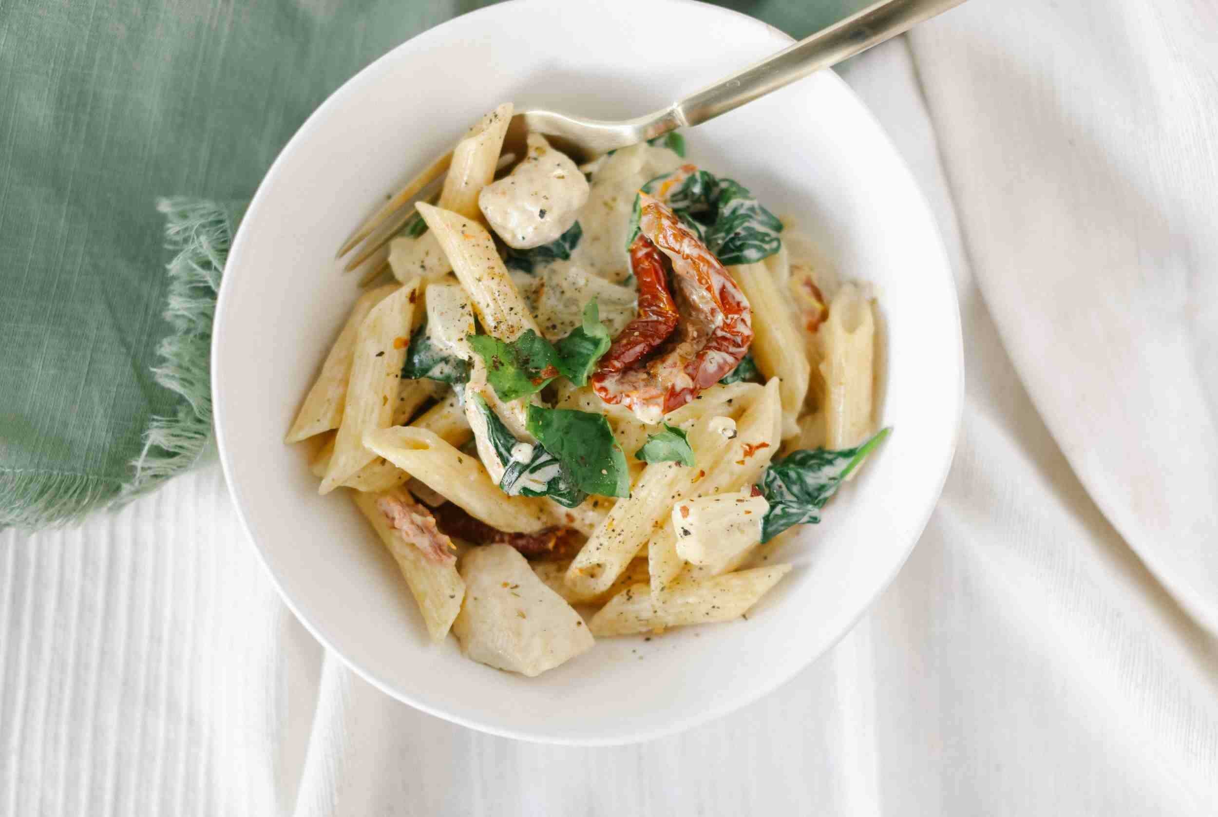 Savory creamy pasta with sun-dried tomatoes, fresh basil, and a blend of cheeses served in a white bowl on a white textured surface.