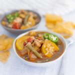 Savory chicken and vegetable soup served in a white bowl with sliced jalapeños, accompanied by crispy tortilla chips and a side of salsa, perfect for a comforting meal.