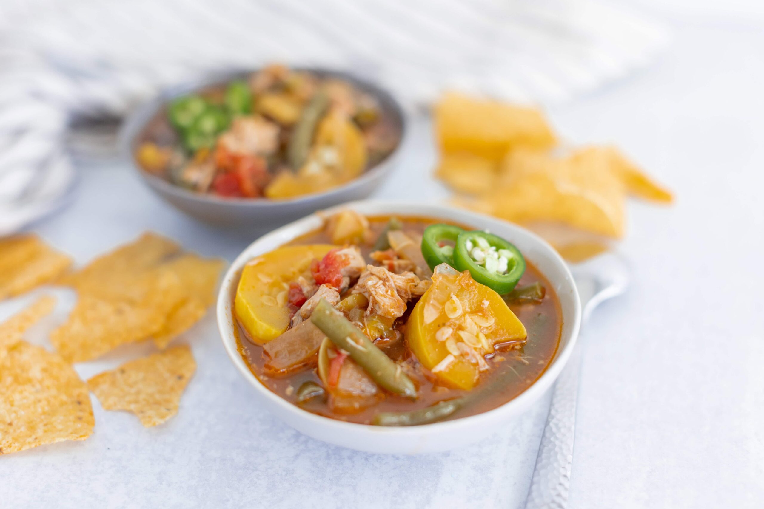Savory chicken and vegetable soup served in a white bowl with sliced jalapeños, accompanied by crispy tortilla chips and a side of salsa, perfect for a comforting meal.