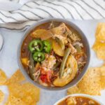 Savory chicken vegetable soup with green chili slices, served with crispy tortilla chips on a white surface, featuring a striped cloth in a bright, inviting kitchen setting.