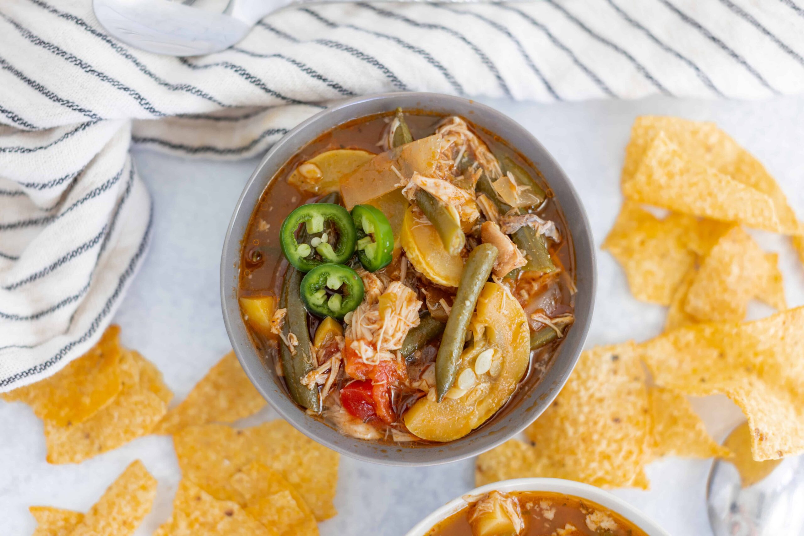 Savory chicken vegetable soup with green chili slices, served with crispy tortilla chips on a white surface, featuring a striped cloth in a bright, inviting kitchen setting.