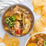 Hearty homemade chicken vegetable soup served in a gray bowl with sliced jalapeños on top, surrounded by crispy tortilla chips, on a white surface with a striped cloth, perfect for comfort food and healthy eating.