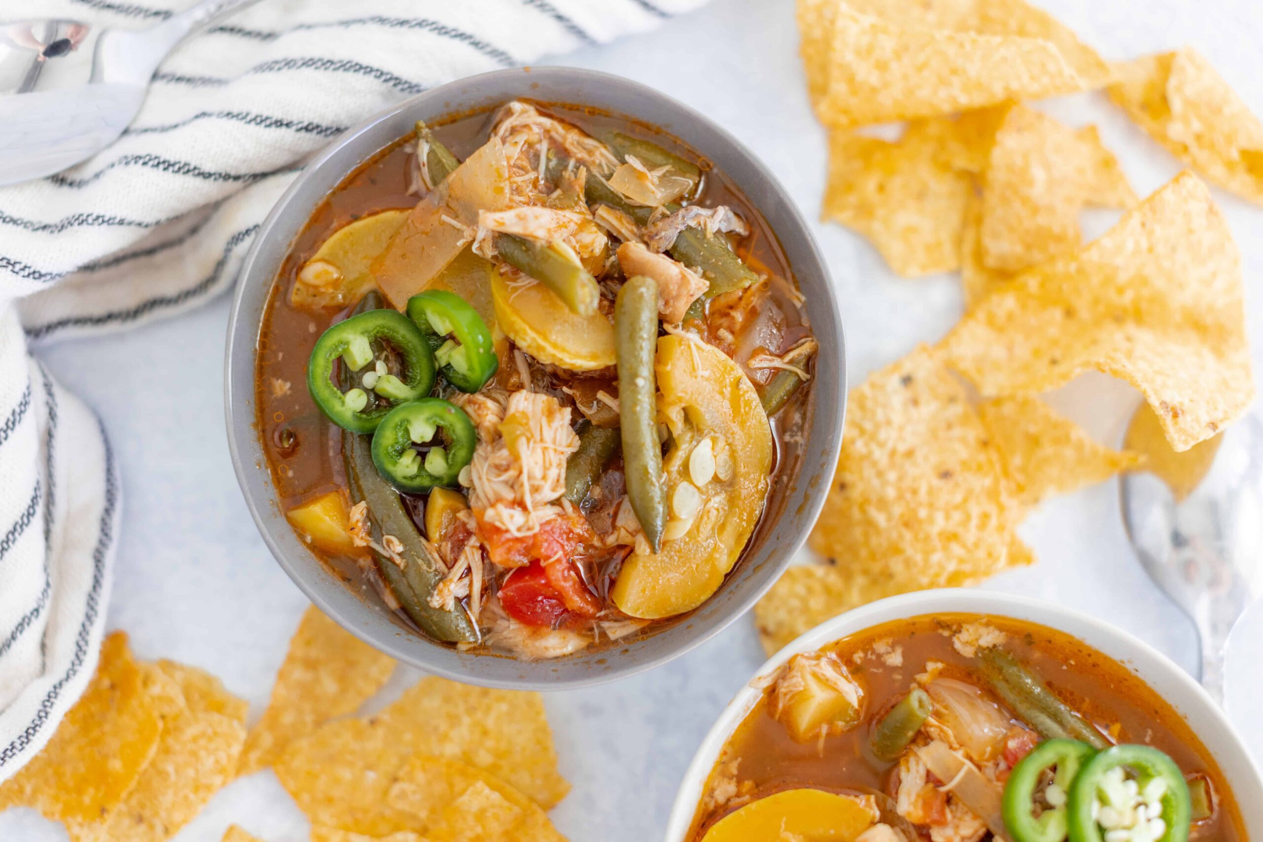Hearty homemade chicken vegetable soup served in a gray bowl with sliced jalapeños on top, surrounded by crispy tortilla chips, on a white surface with a striped cloth, perfect for comfort food and healthy eating.