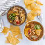 Hearty vegetable chicken soup served in two bowls with slices of jalapeños on top, accompanied by crispy tortilla chips, on a white surface with a striped cloth for a comforting meal.