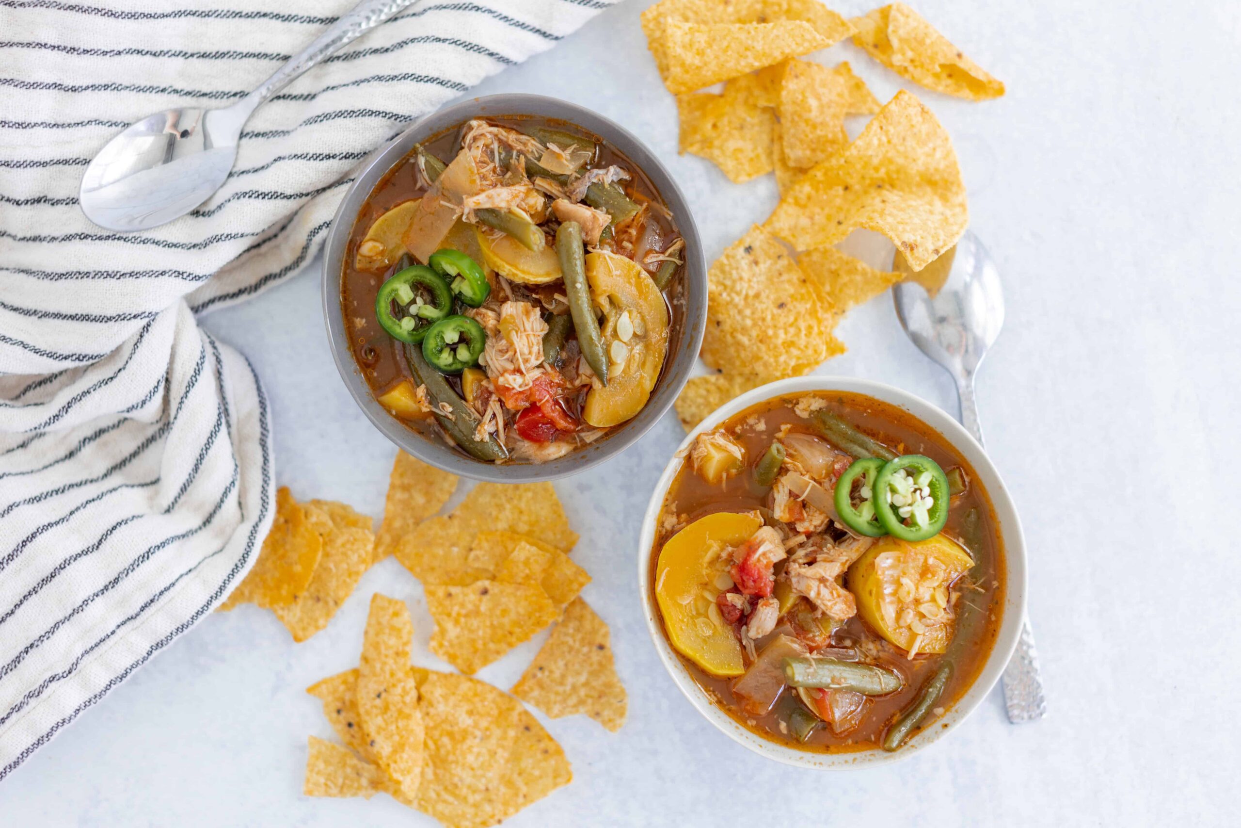 Hearty vegetable chicken soup served in two bowls with slices of jalapeños on top, accompanied by crispy tortilla chips, on a white surface with a striped cloth for a comforting meal.