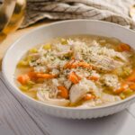 Comforting chicken and vegetable soup in a white bowl, featuring shredded chicken, carrots, celery, and herbs, served on a rustic wooden table.