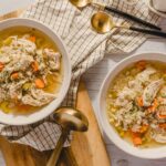 Juicy chicken and vegetable soup with carrots, celery, and herbs served in white bowls, perfect for comforting homemade meals; casual dining setup with gold spoons and checkered napkin.