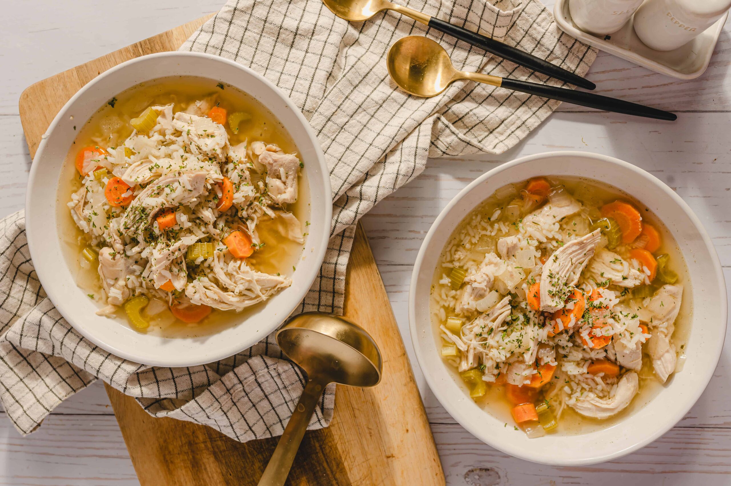 Juicy chicken and vegetable soup with carrots, celery, and herbs served in white bowls, perfect for comforting homemade meals; casual dining setup with gold spoons and checkered napkin.