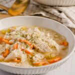 Savory chicken and vegetable soup in a white bowl, featuring shredded chicken, carrots, celery, and herbs, served with a spoon on a rustic table.