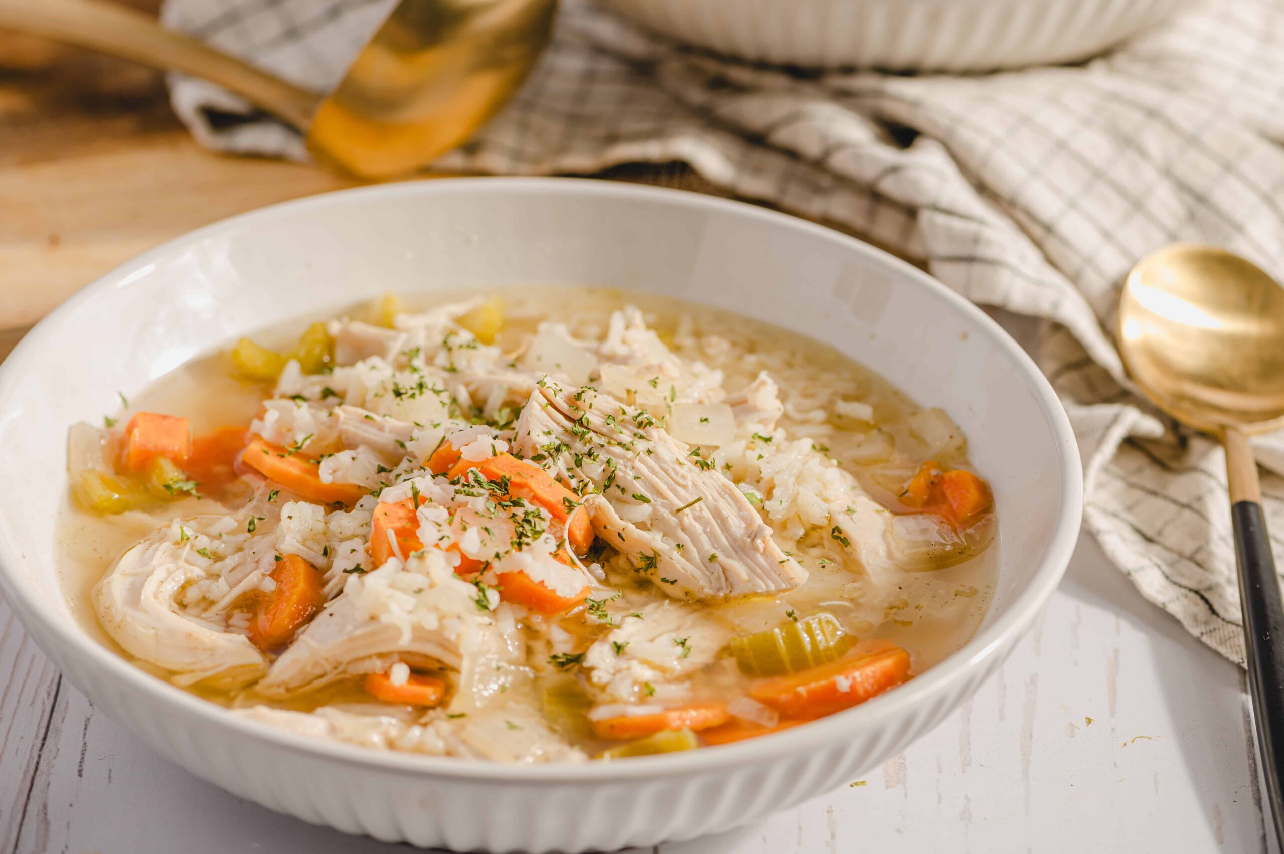 Savory chicken and vegetable soup in a white bowl, featuring shredded chicken, carrots, celery, and herbs, served with a spoon on a rustic table.