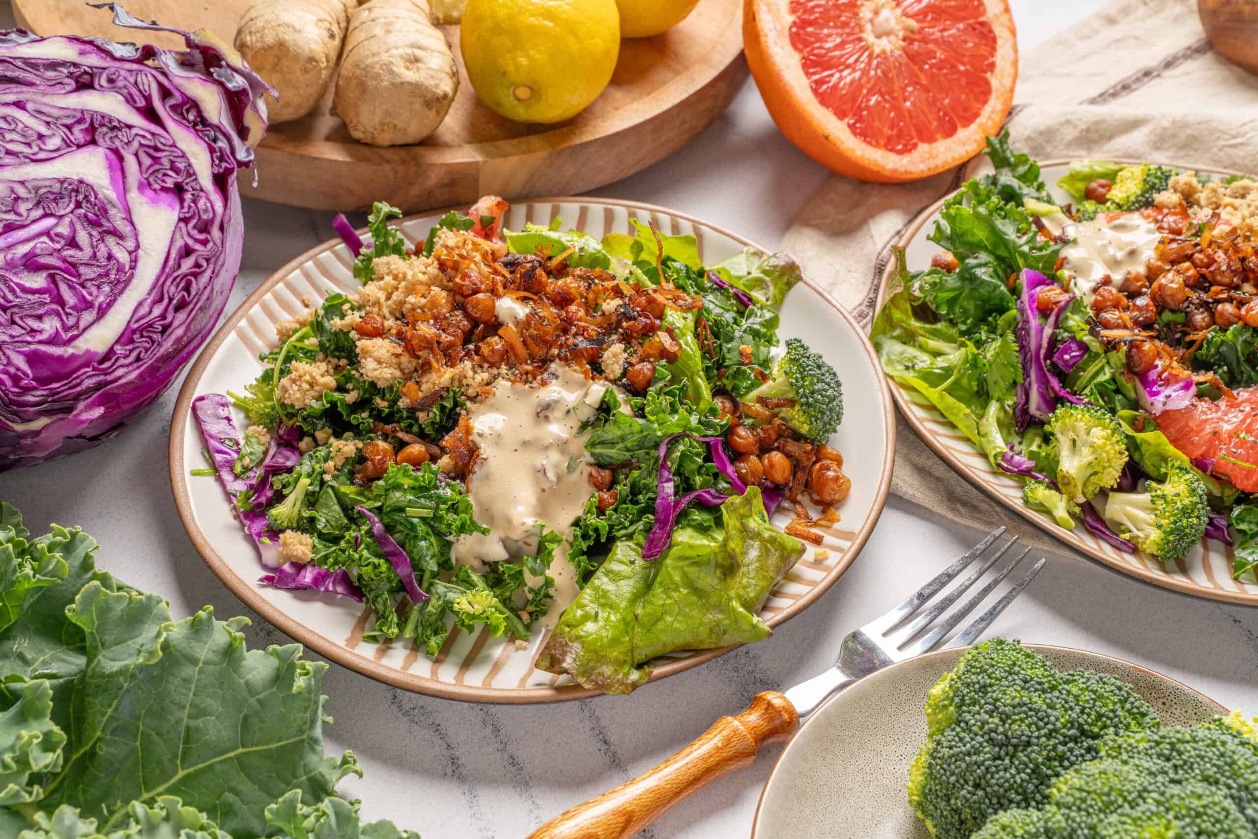 Fresh vegetable salad with broccoli, cabbage, cherry tomatoes, and a creamy dressing, served on decorative plates, with fresh ingredients like grapefruit, ginger, and garlic surrounding it.