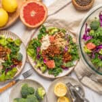 Fresh kale and broccoli salad with colorful vegetables, citrus, and crunchy toppings on striped plates, surrounded by lemons, ginger, and herbs for healthy eating recipes.