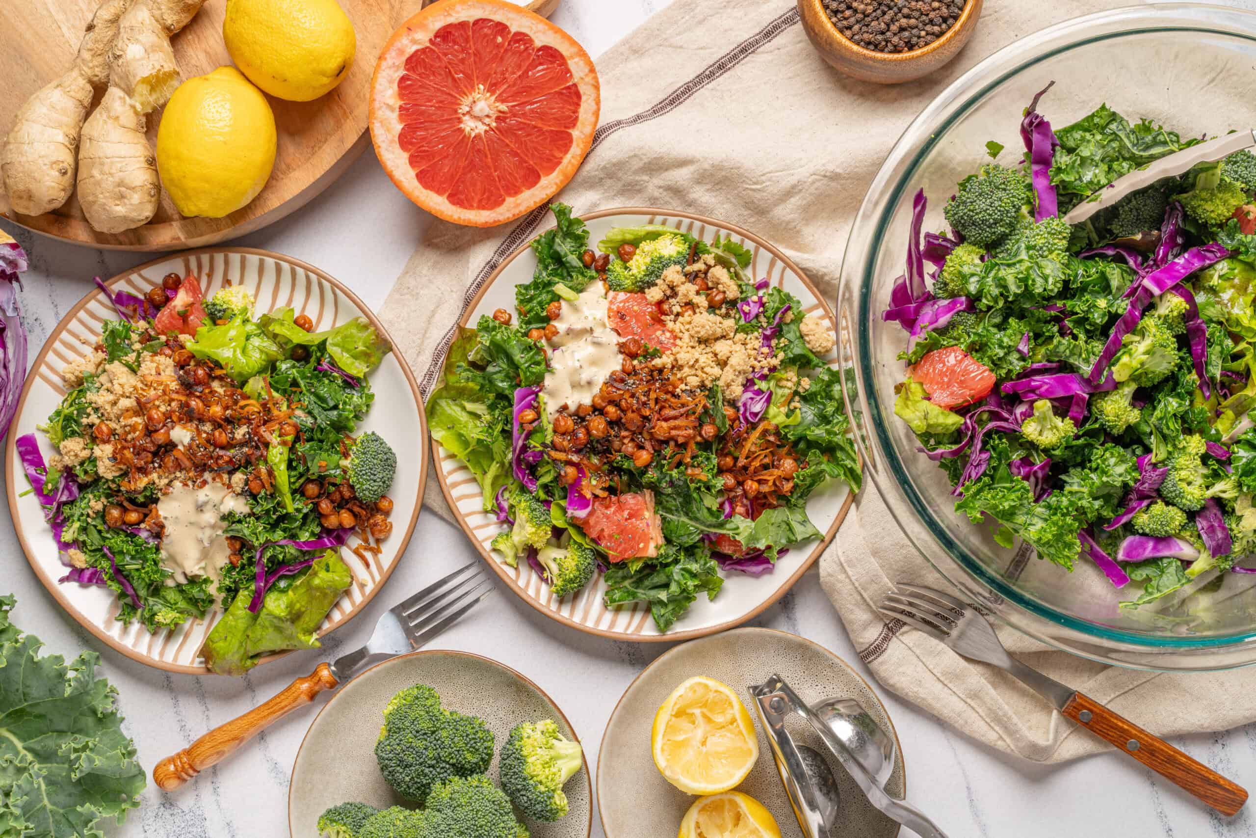 Fresh kale and broccoli salad with colorful vegetables, citrus, and crunchy toppings on striped plates, surrounded by lemons, ginger, and herbs for healthy eating recipes.