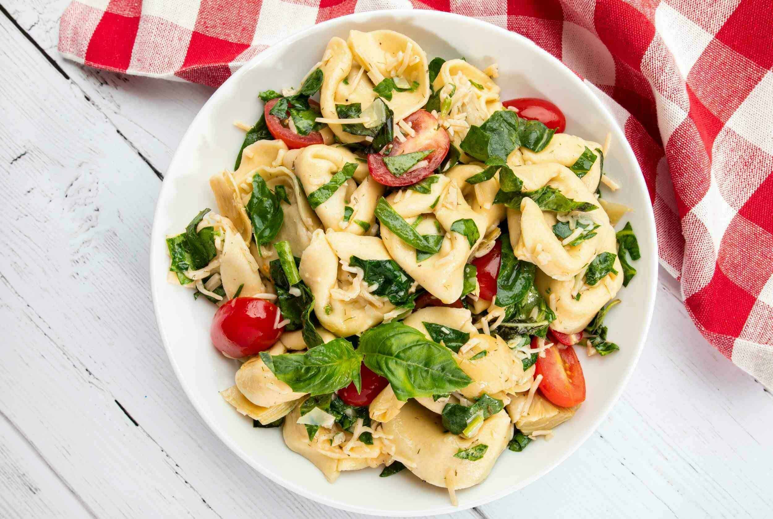 Fresh tortellini pasta salad with cherry tomatoes, basil, and shredded cheese on a white plate, served with a red and white checkered cloth in the background.