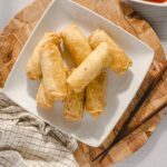 Crispy spring rolls on a white plate with chopsticks, served on a wooden platter, perfect for appetizer or snack, featured on Hey Recipe website.