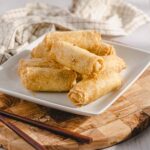 Crispy spring rolls with golden, crunchy exterior served on a white platter with a side of sweet chili dipping sauce, garnished with chopsticks on a rustic wooden board.